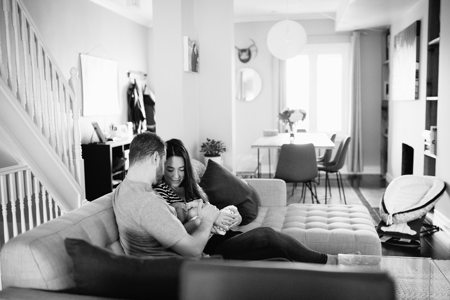 family of 3, new parents cuddle on couch, mom holding 2 week old baby girl covered with black and white swaddle, dad embracing wife and daughter - York Region Lifestyle Photos