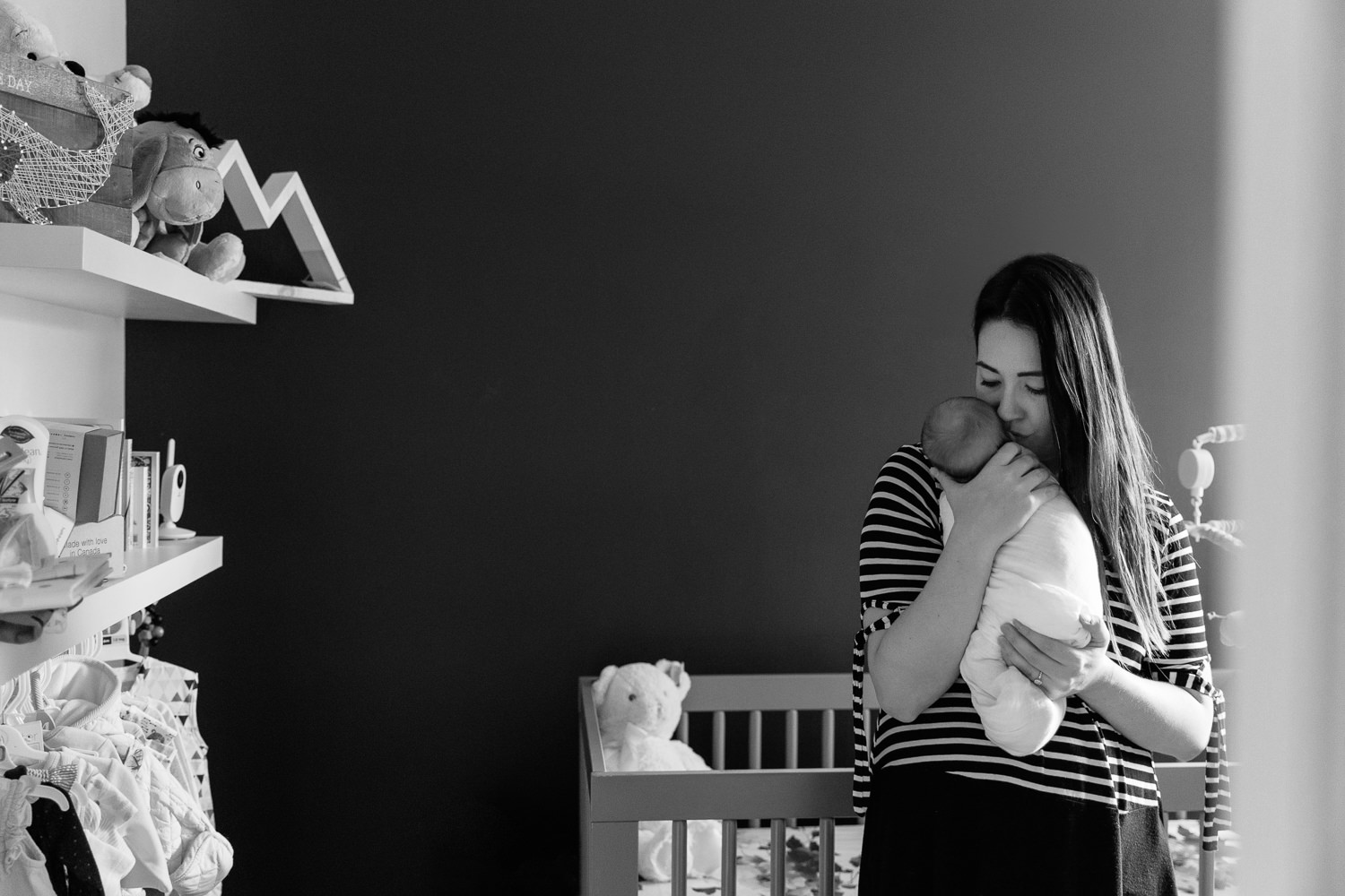 new mom standing in nursery in front of crib holding 2 week old sleeping baby girl wrapped in white swaddle to her chest, mother's eyes closed - Markham Lifestyle Photos