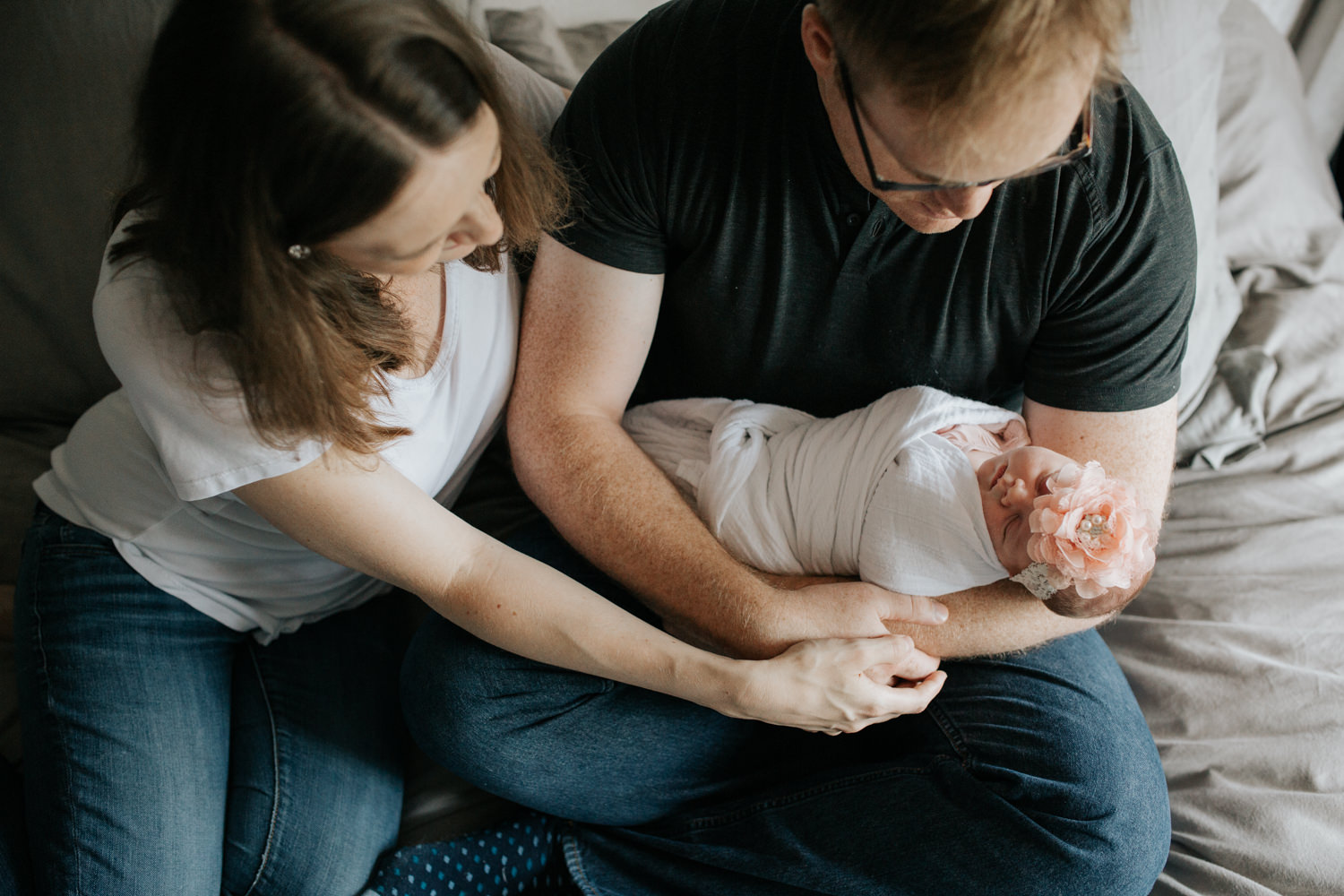 family of 3, new parents sitting on bed, dad holding 2 week old swaddled baby girl as mom snuggles next to husband smiling at daughter - Stouffville Lifestyle Photography
