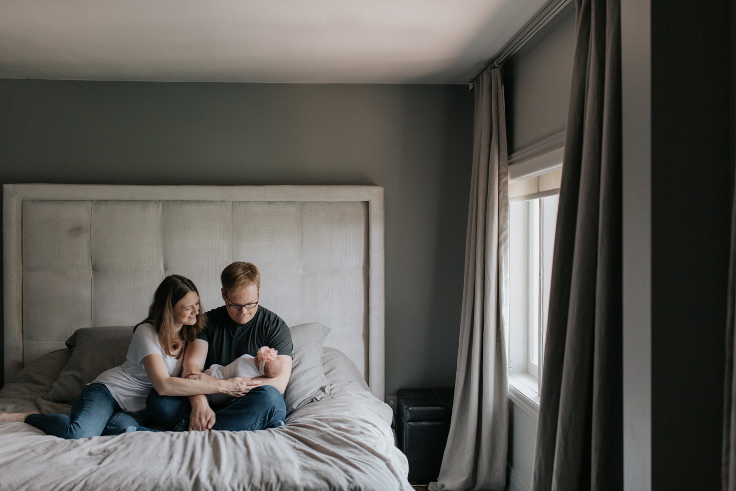 family of 3, new parents sitting on bed, dad holding 2 week old swaddled baby girl as mom snuggles next to husband smiling at daughter - Newmarket Lifestyle Photography
