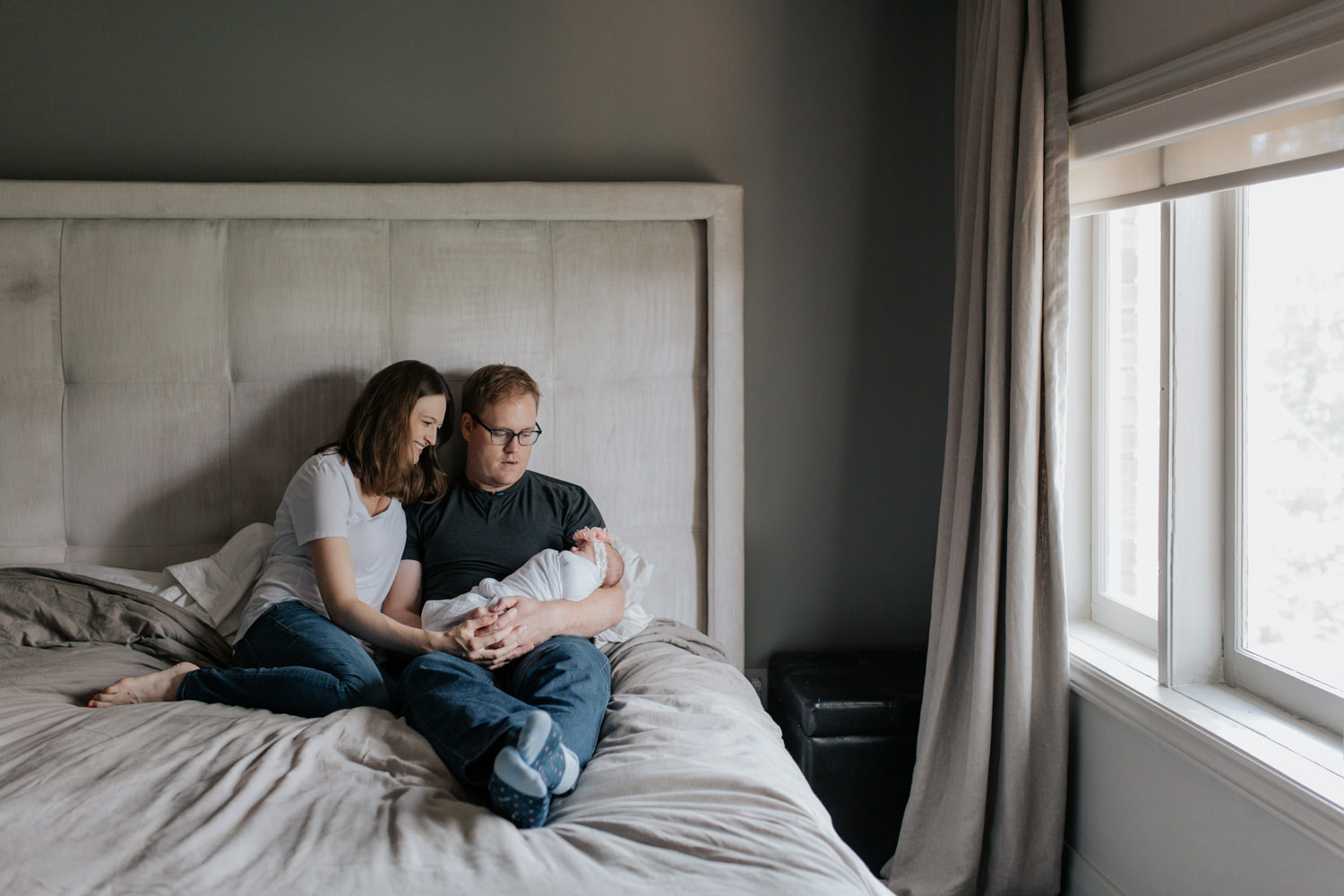 family of 3, new parents sitting on bed, dad holding 2 week old swaddled baby girl as mom snuggles next to husband smiling at daughter - Uxbridge Lifestyle Photography