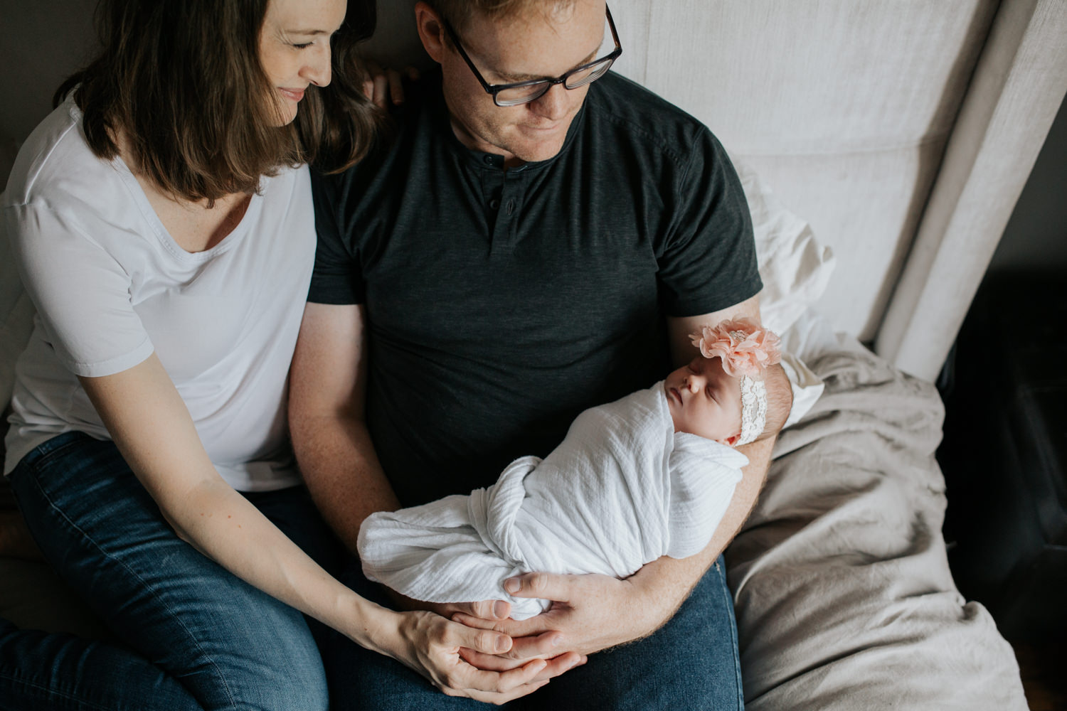 family of 3, new parents sitting on bed, dad holding 2 week old swaddled baby girl as mom snuggles next to husband smiling at daughter - GTA Lifestyle Photography