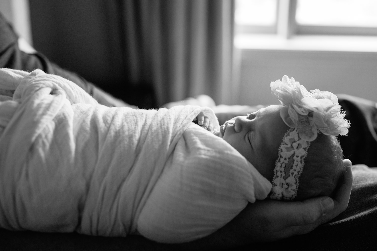 2 week old baby girl in white swaddle withflower headband lying asleep in dad's arms as he sits on bed with legs stretched out -Newmarket In-Home Photography