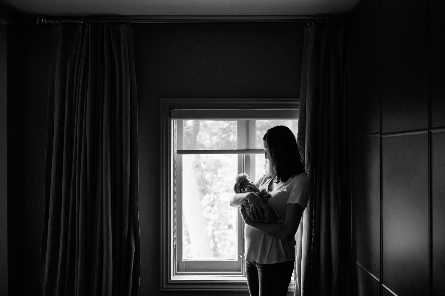 new mom standing in front of window in dark room, holding and looking at 2 week old baby girl in her arms - Uxbridge In-Home Photos