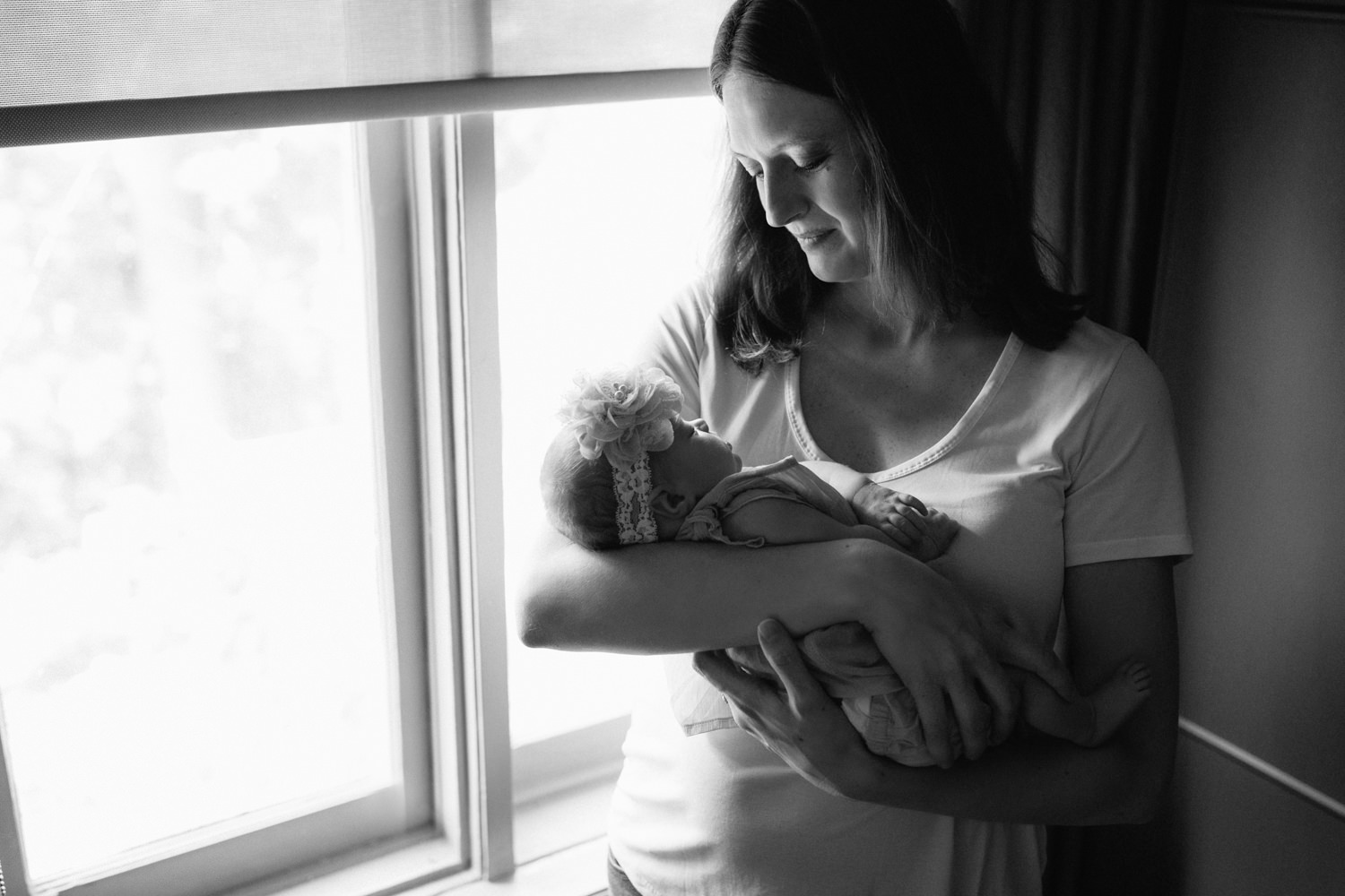 new mom in white t-shirt standing in front of window in dark room, holding and looking at 2 week old baby girl in her arms - et in the air - GTA In-Home Photos