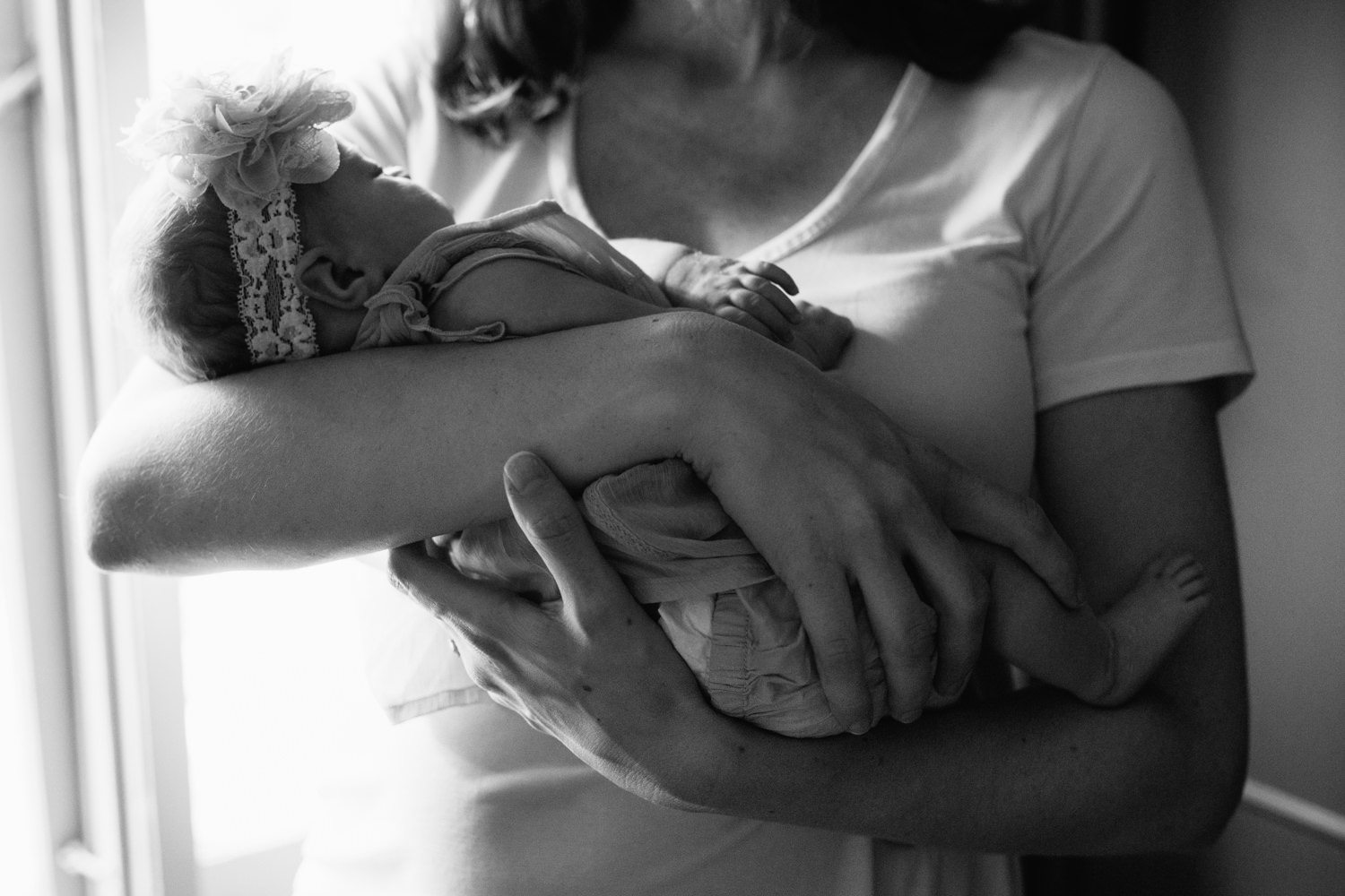 new mom in white t-shirt standing in front of window in dark room, holding 2 week old baby girl with flower headband on in her arms - York Region In-Home Photos