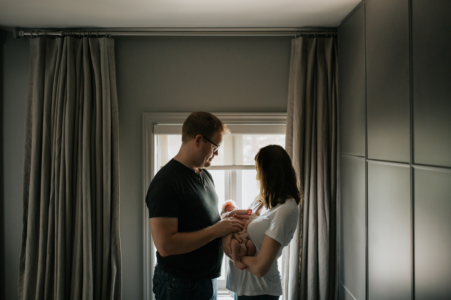 new parents standing in front of window, mom holding 2 week old baby girl in her arms, dad standing in front of wife hand on daughter - Newmarket Lifestyle Photos