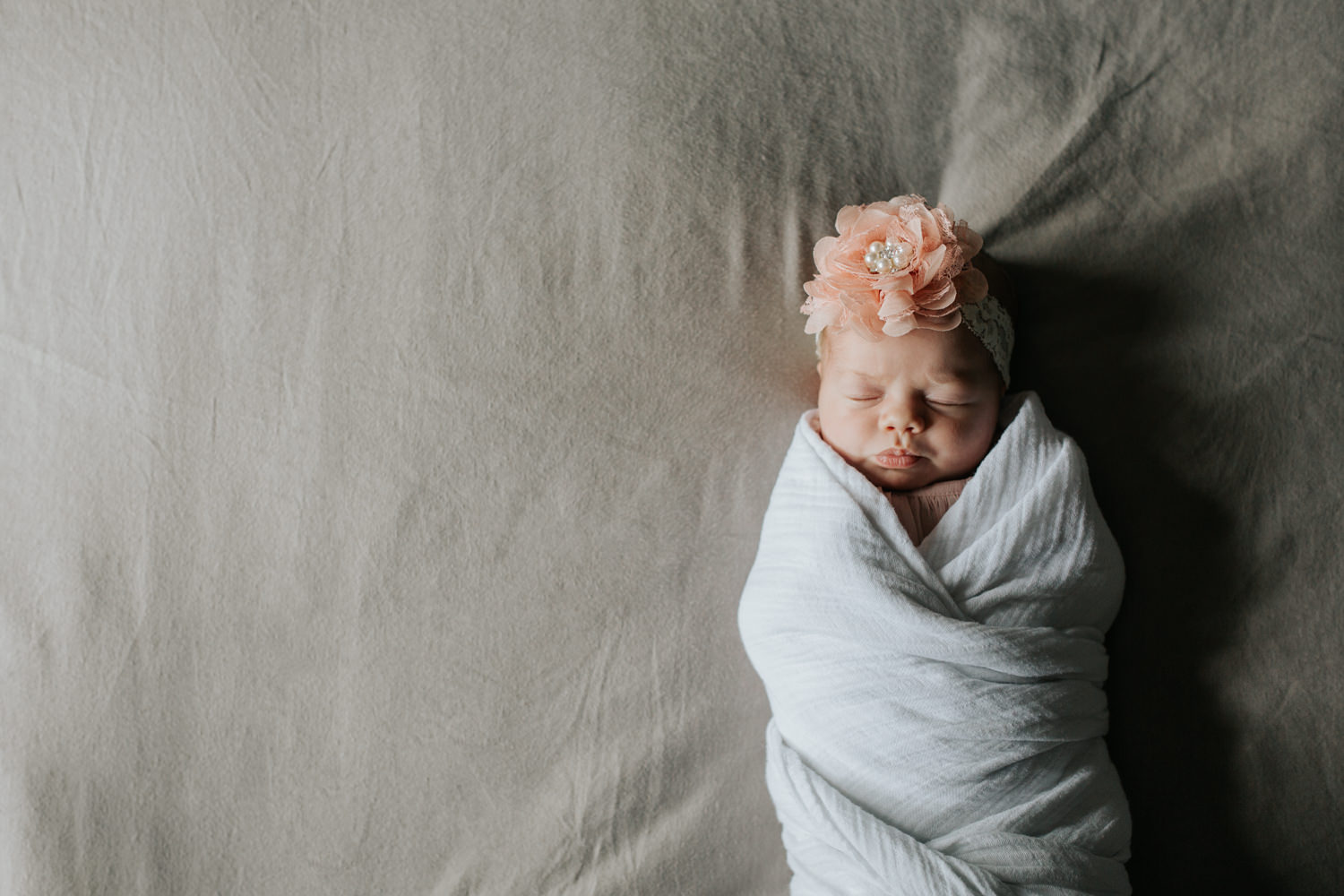 2 week old baby girl wrapped in white swaddle wearing pink flower headband fast asleep lying on master bed - Stouffville Lifestyle Photography