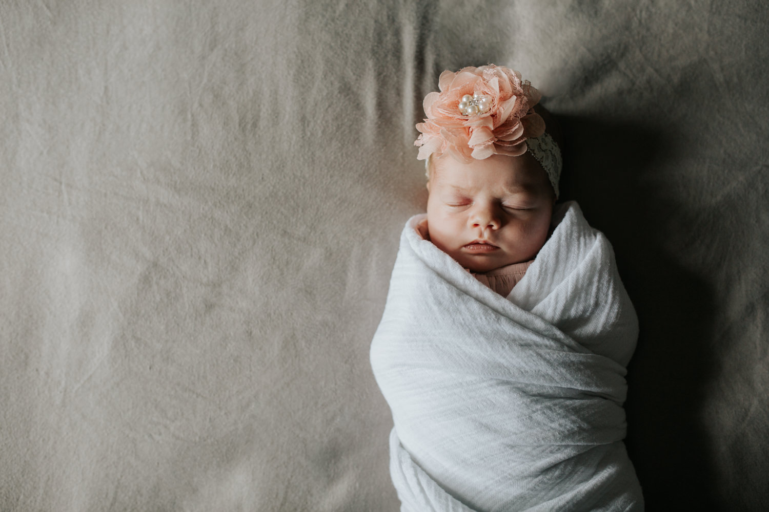 2 week old baby girl wrapped in white swaddle wearing pink flower headband fast asleep lying on master bed - Newmarket Lifestyle Photography