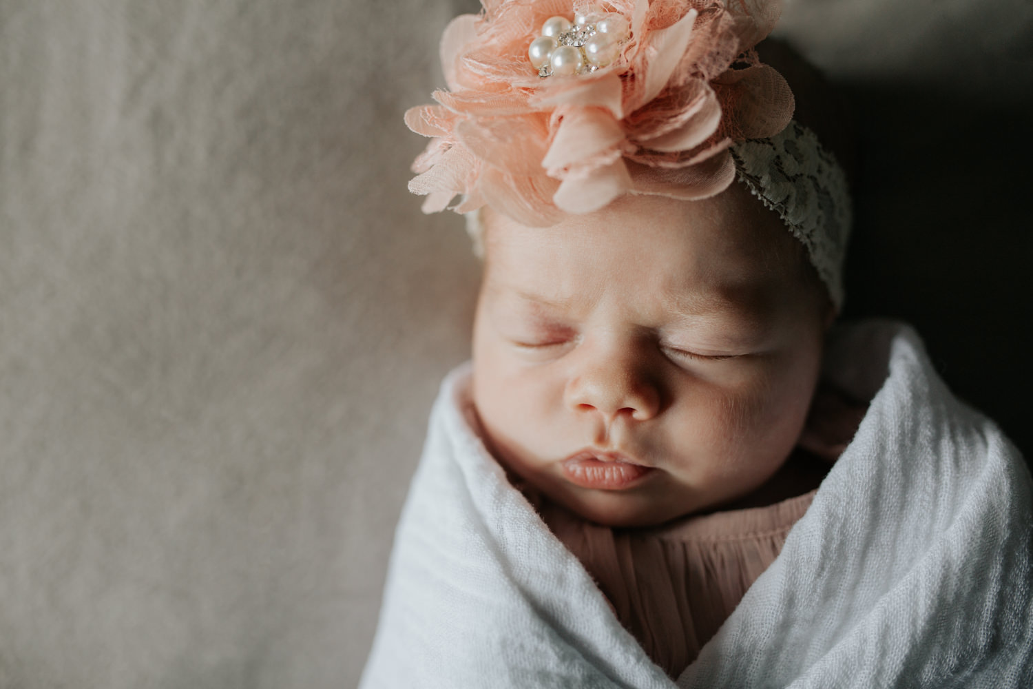 2 week old baby girl wrapped in white swaddle wearing pink flower headband fast asleep lying on master bed, lips pursed - Markham Lifestyle Photography