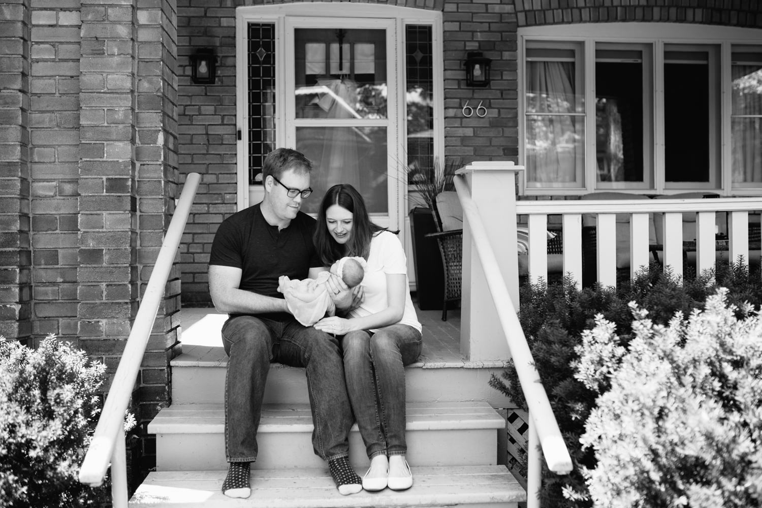 family of 3 sitting on front porch on summer day, dad holding 2 week old baby girl, mom leaning in next to husband smiling at daughter - Barrie Lifestyle Photography