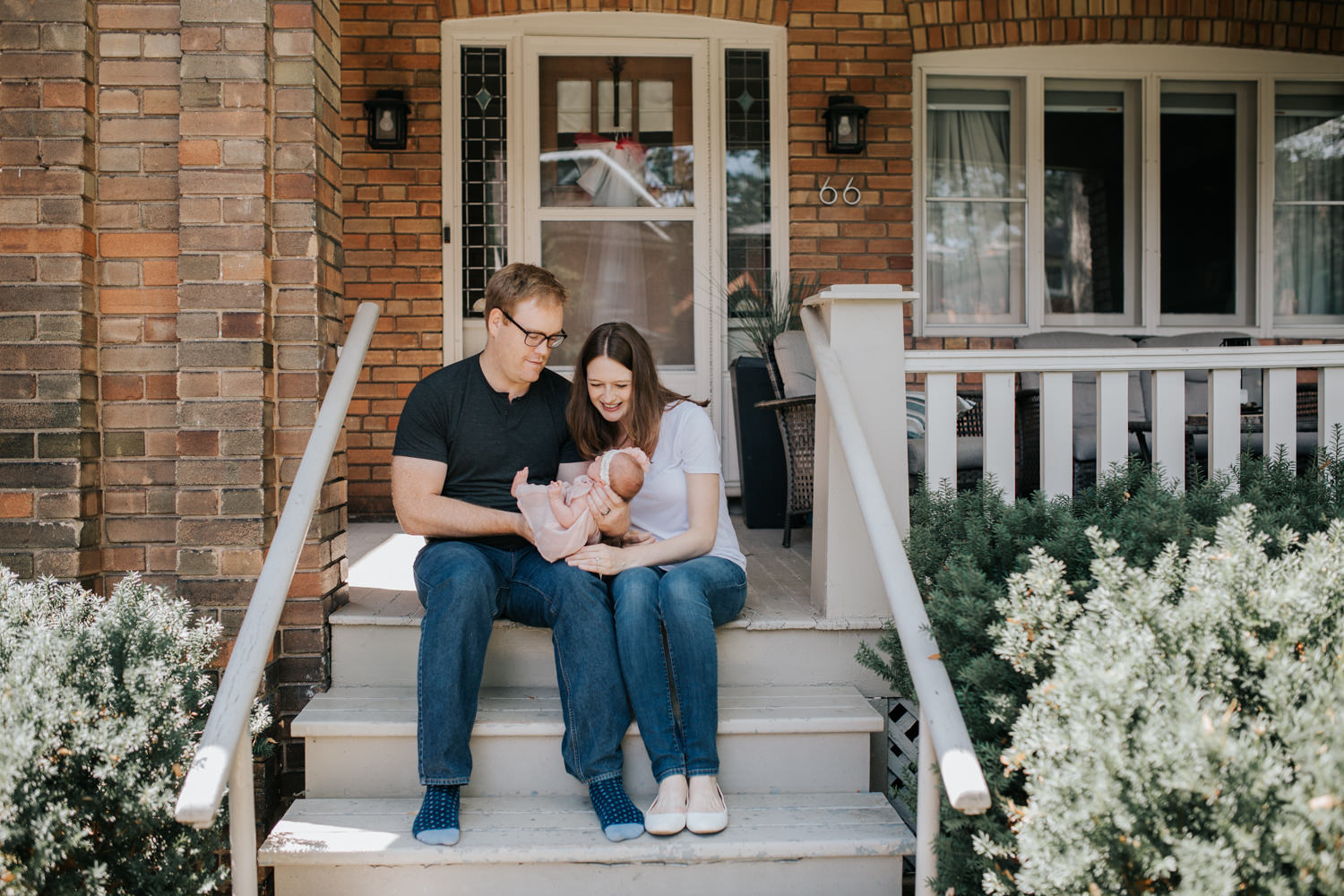 family of 3 sitting on front porch on summer day, dad holding 2 week old baby girl, mom leaning in next to husband smiling at daughter - GTA Lifestyle Photography