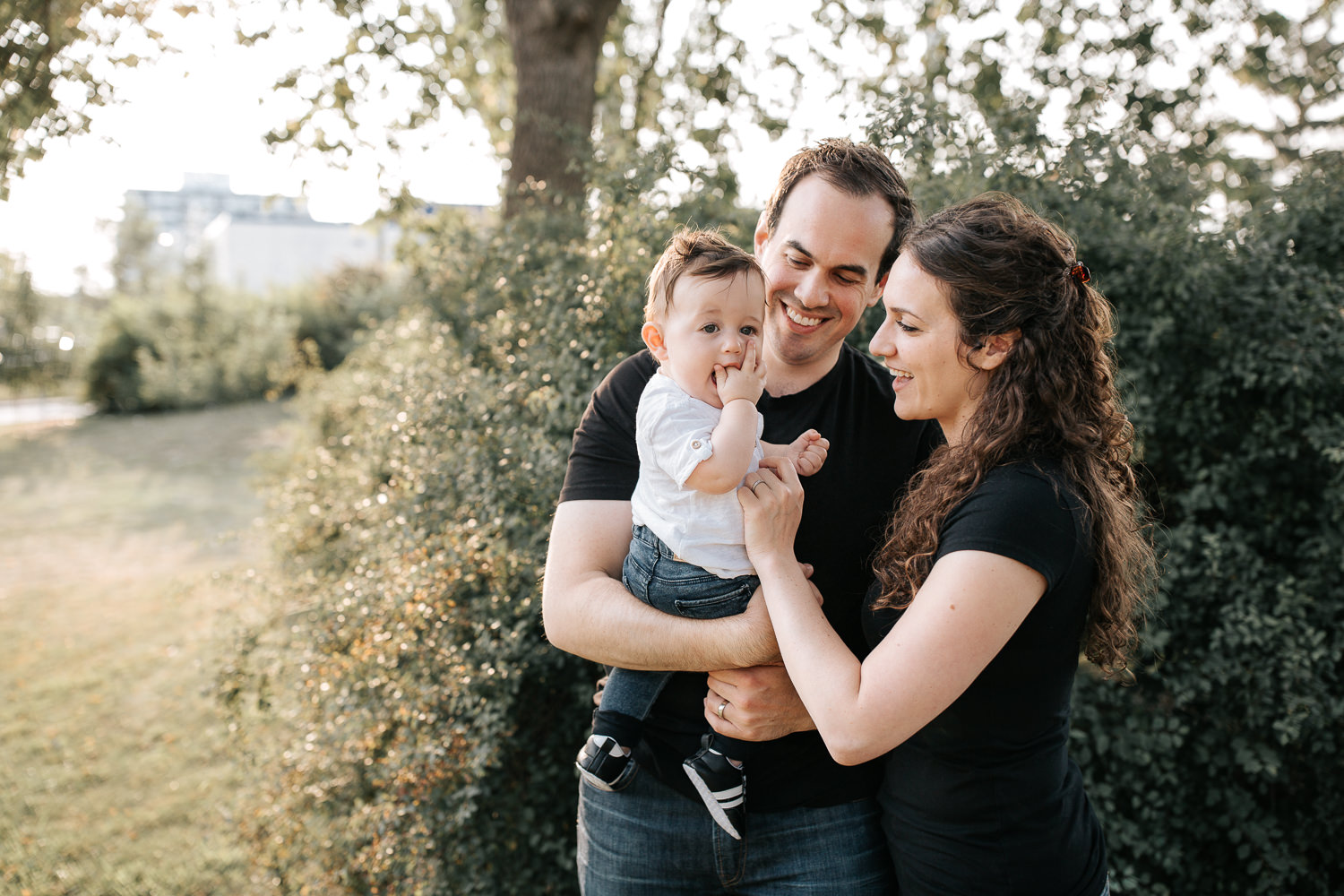 family of 3, smiling parents standing in park holding 9 month old baby boy with brown hair, chewing on hand - Newmarket Lifestyle Photography