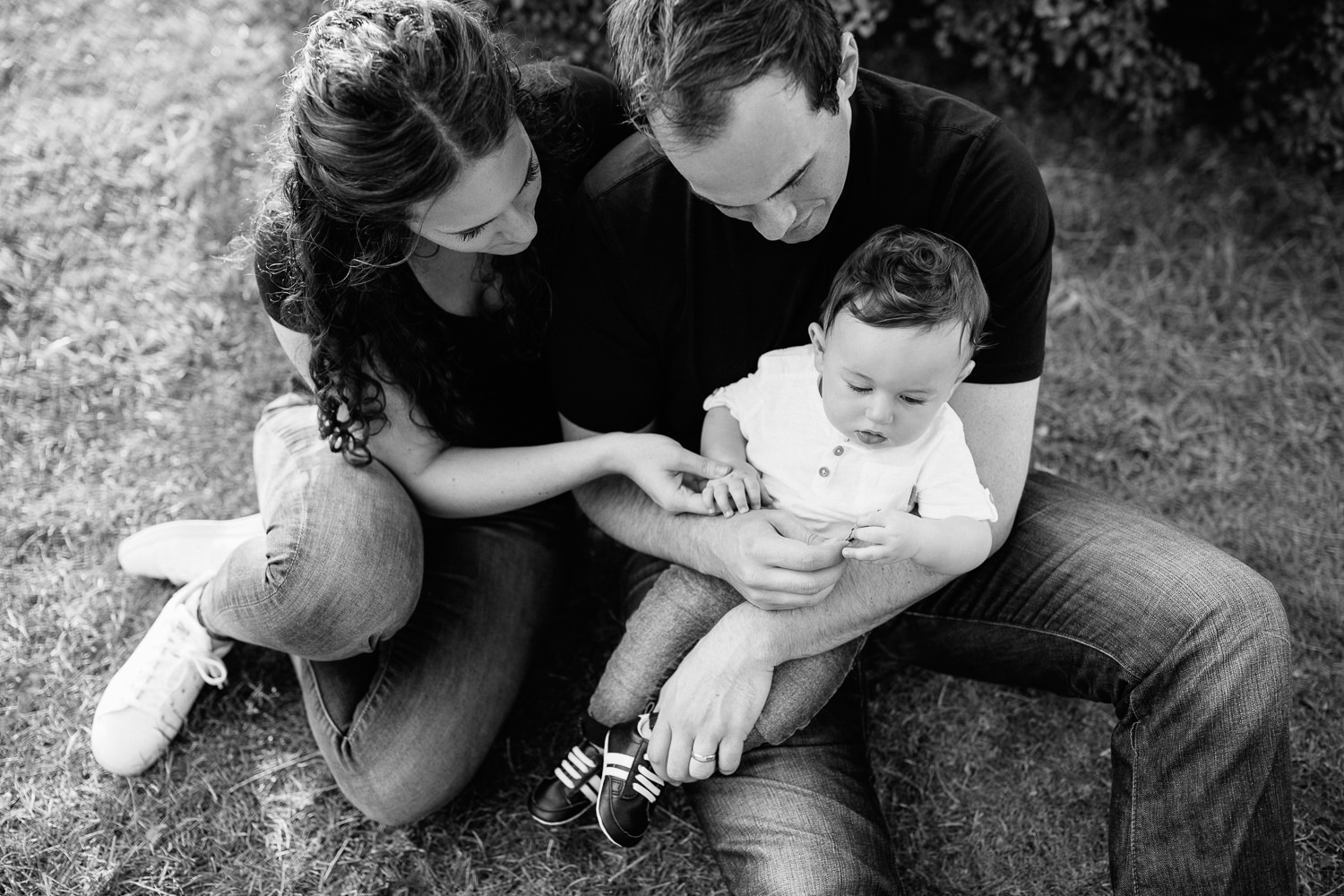 family of 3 sitting on ground in park, 9 month old baby boy with dark hair sitting in dad's lap, mom snuggled next to them holding son's hand - York Region Lifestyle Photography