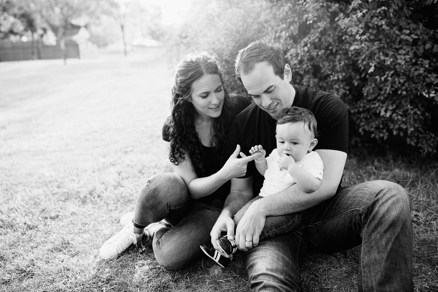 family of 3 sitting on ground in park, 9 month old baby boy with dark hair sitting in dad's lap, mom snuggled next to them holding son's hand - Barrie Lifestyle Photography