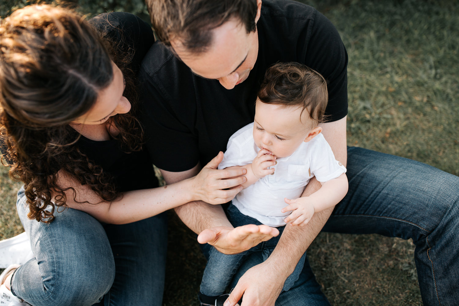 family of 3 sitting on ground in park, 9 month old baby boy with dark hair sitting in dad's lap, mom snuggled next to them, hand on son - GTA Lifestyle Photography