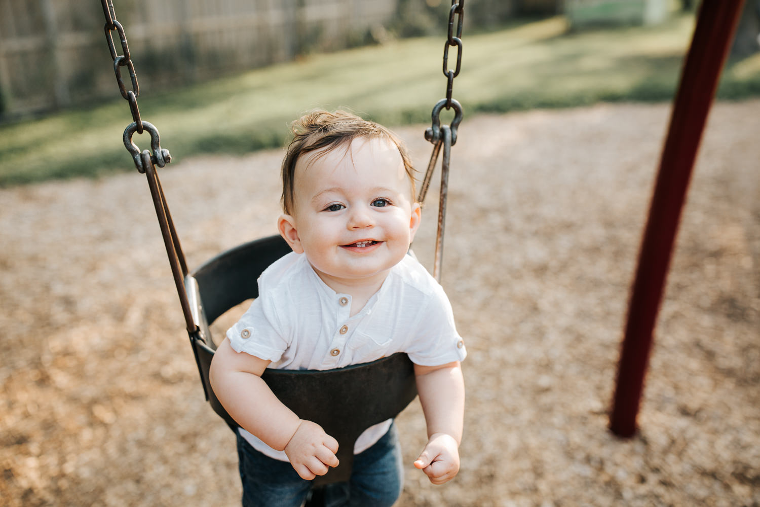9 month old baby boy with dark brown hair wearing white t-shirt and jeans smiling looking at camera as he sits in swing at park - Newmarket Golden Hour Photography