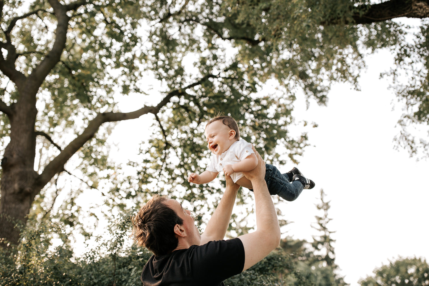 9 month old baby boy with dark brown hair wearing white t-shirt and jeans laughing as dad throws him in air under trees in park - Stouffville Lifestyle Photos