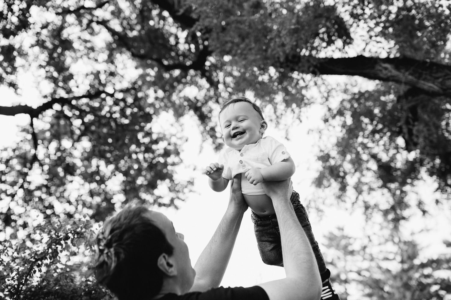 9 month old baby boy with dark brown hair wearing white t-shirt and jeans laughing as dad throws him in air under trees in park - Barrie Lifestyle Photos