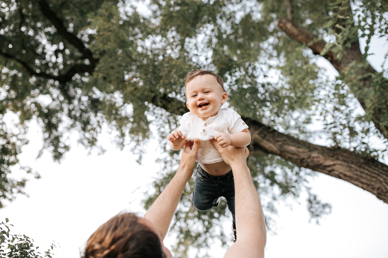 9 month old baby boy with dark brown hair wearing white t-shirt and jeans laughing as dad throws him in air under trees in park - GTA Lifestyle Photos