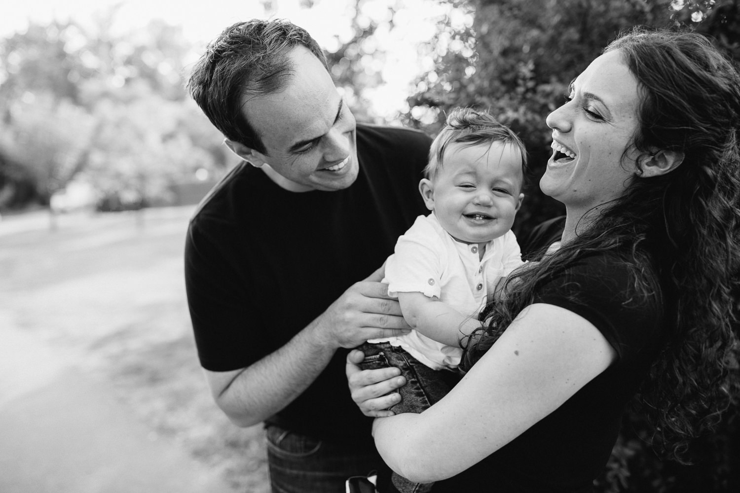 family of 3, smiling parents standing in park holding 9 month old baby, mom and dad laughing while baby cries - Stouffville Lifestyle Photography