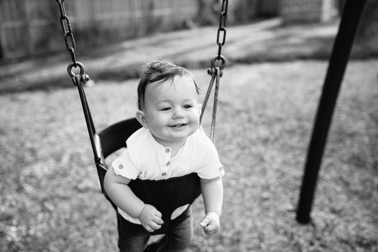 9 month old baby boy with dark brown hair wearing white t-shirt and jeans smiling as he sits in swing at park - Barrie Golden Hour Photography