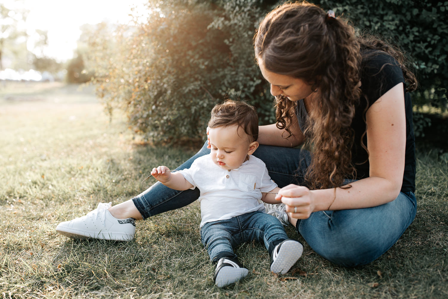 9 month old baby boy with dark brown hair wearing white t-shirt, jeans and sneakers sitting on ground playing with grass at park, mom sitting next to son - GTA Golden Hour Photography