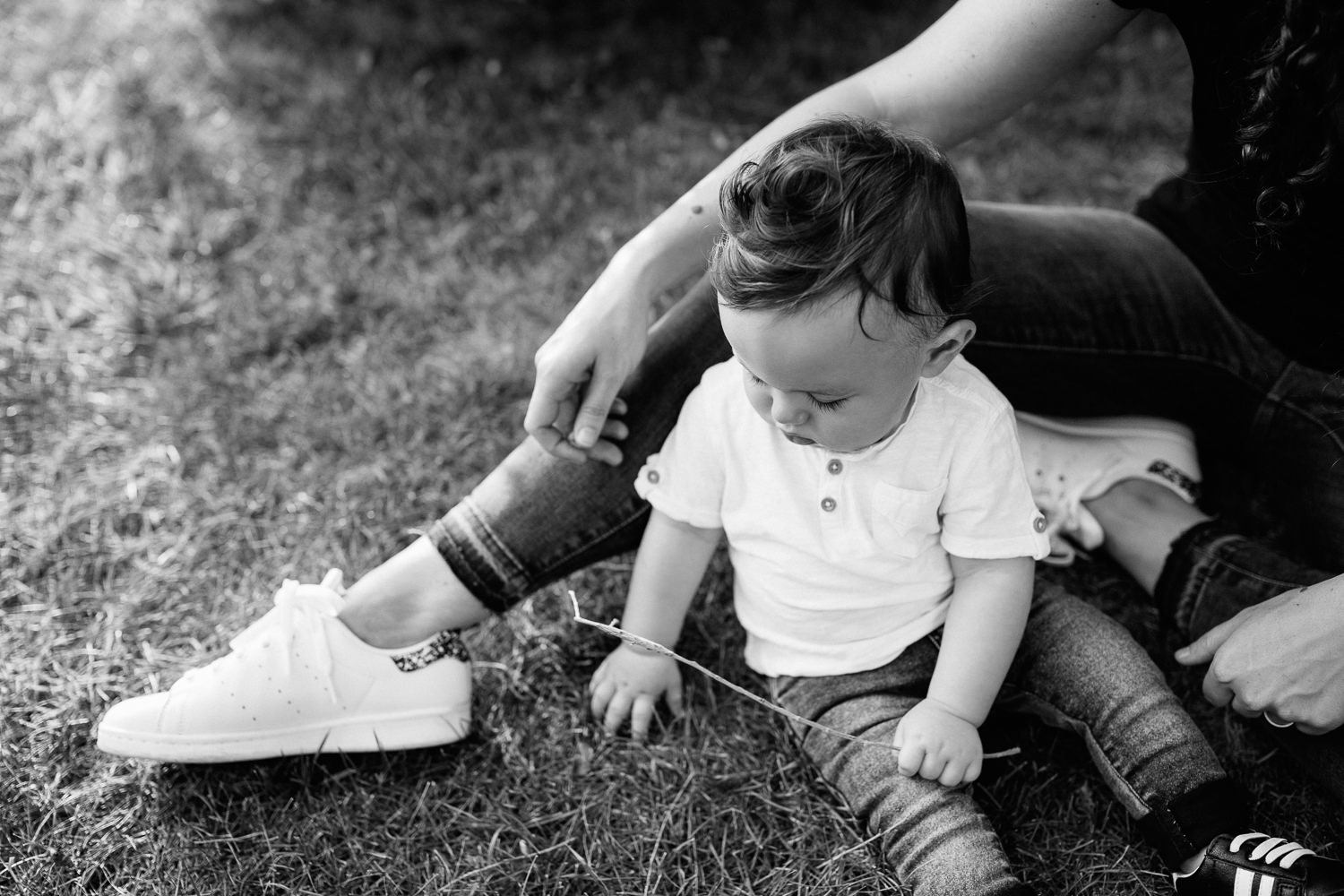 York9 month old baby boy with dark brown hair wearing white t-shirt, jeans and sneakers sitting on ground playing with grass at park, mom sitting next to son -  Region Golden Hour Photography