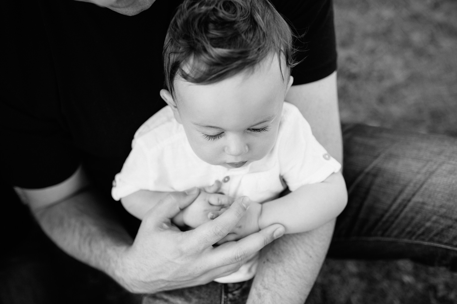 9 month old baby boy wearing white t-shirt and jeans sitting in dad's lap with hands folded, looking down - Stouffville Golden Hour Photos