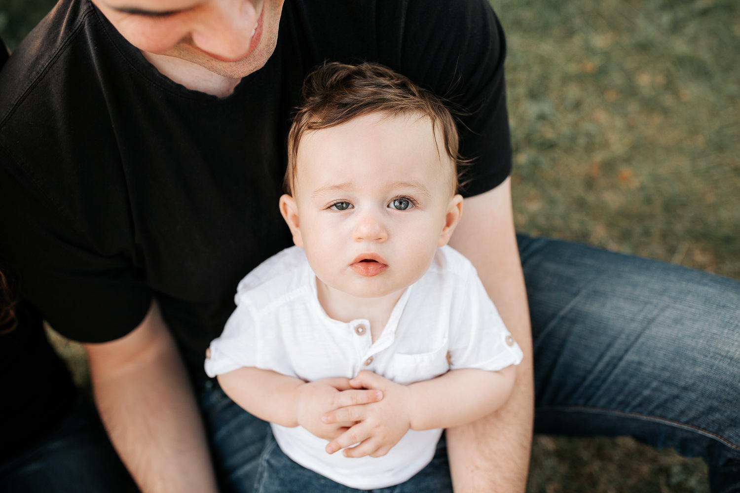 9 month old baby boy with blue eyes wearing white t-shirt and jeans sitting in dad's lap with hands folded, looking up at camera - Newmarket Golden Hour Photos