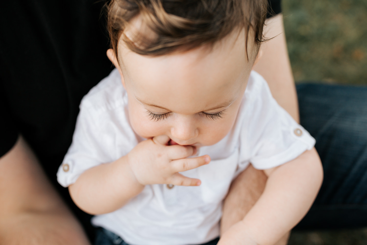 9 month old baby boy wearing white t-shirt and jeans sitting in dad's lap with hands folded, looking down, close up of eyelashes - Markham Golden Hour Photos