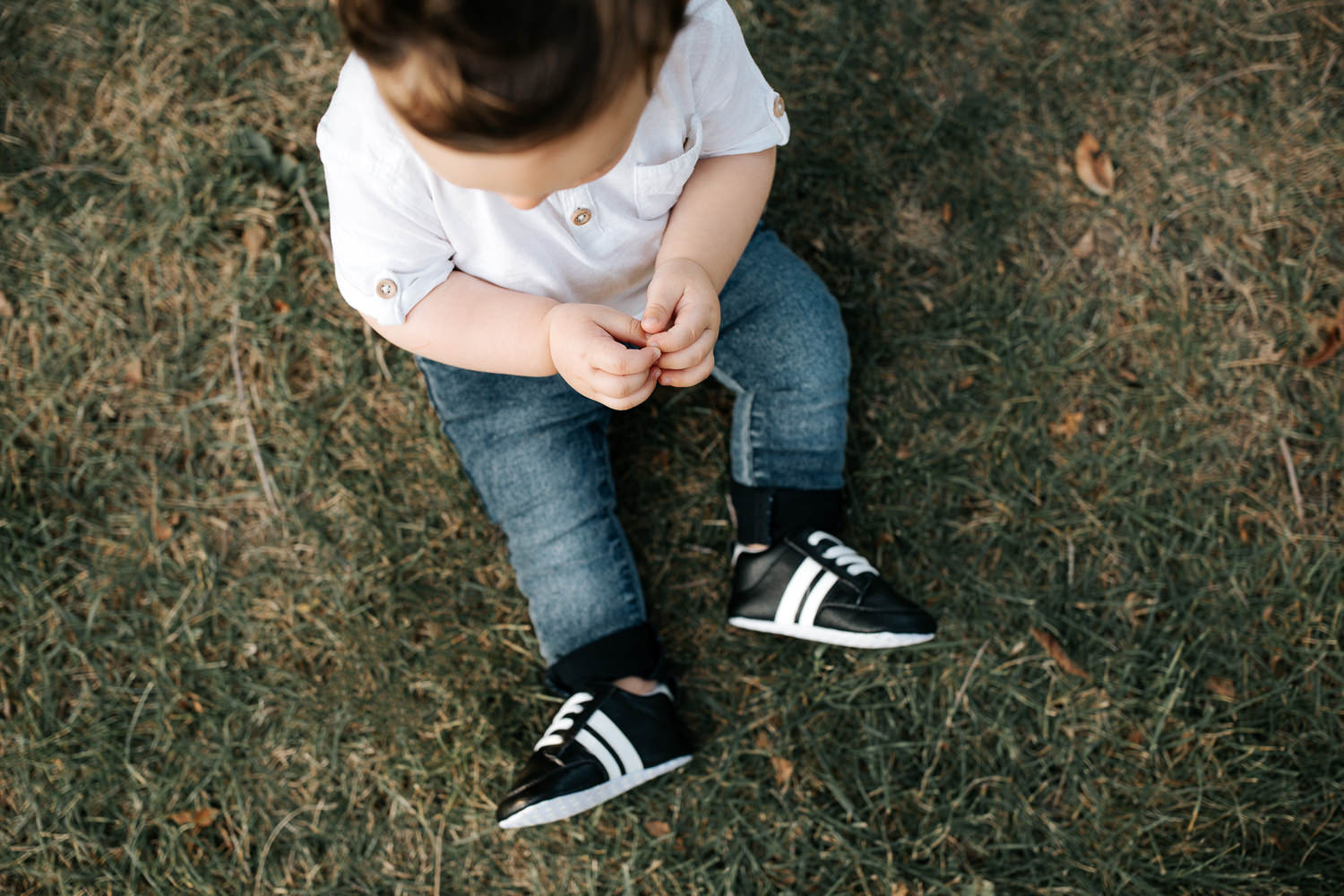 9 month old baby boy wearing white t-shirt, jeans and sneakers sitting on grass at park in front of greenery, close up of clasped hands and feet - GTA Golden Hour Photos