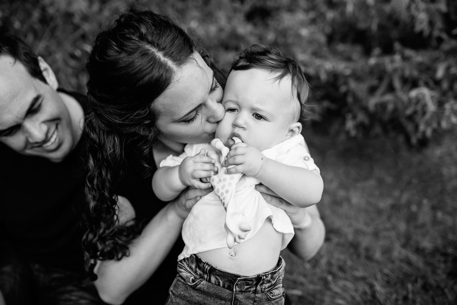 mom holding up 9 month old baby boy with dark hair chewing on Sophie the giraffe, mother kissing son on cheek, dad sitting behind smiling - Stouffville Lifestyle Photography