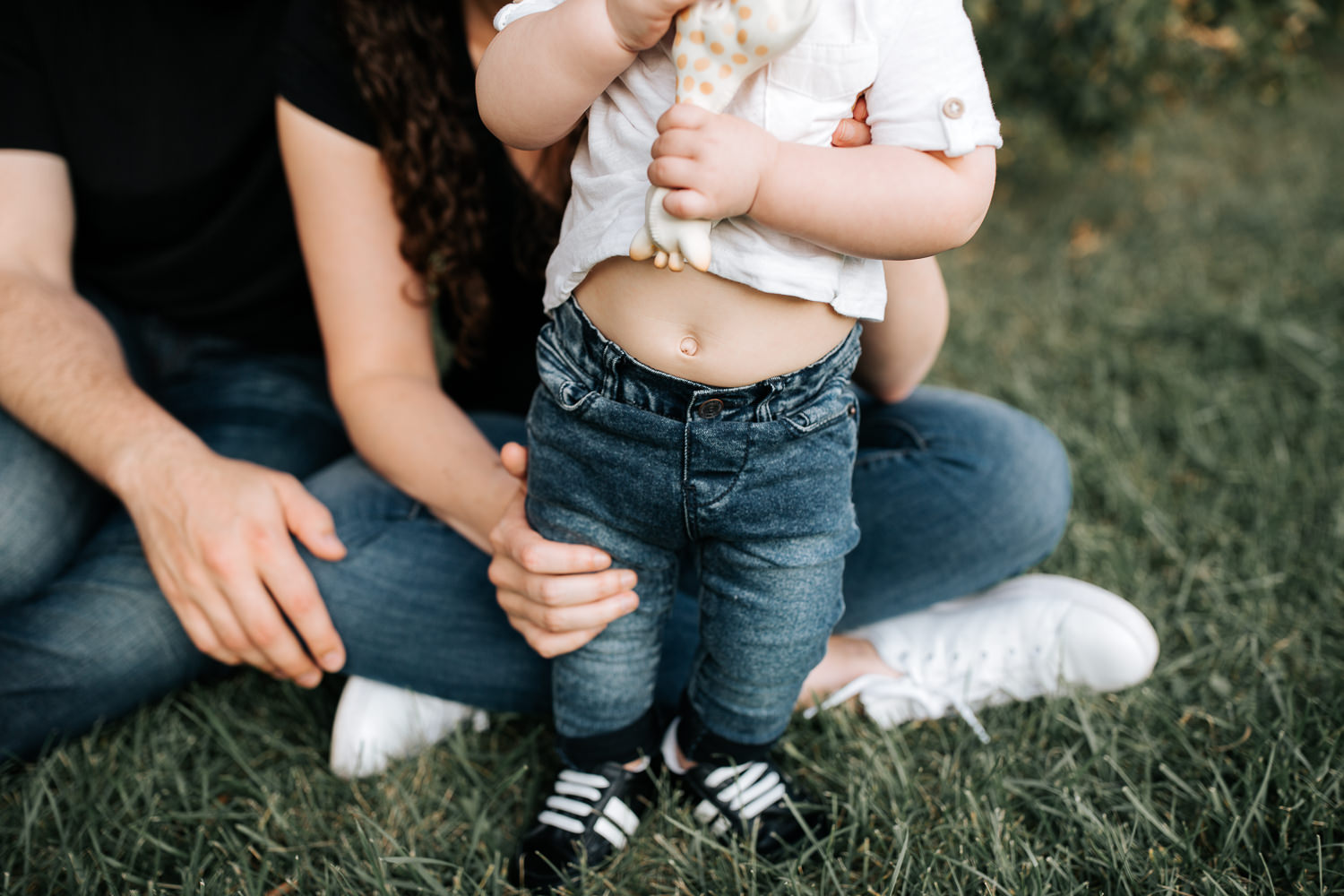 9 month old baby boy in white t-shirt, jeans and sneakers standing holding Sophie the giraffe, mom and dad sitting behind son, close up of bellybutton - Newmarket Lifestyle Photography