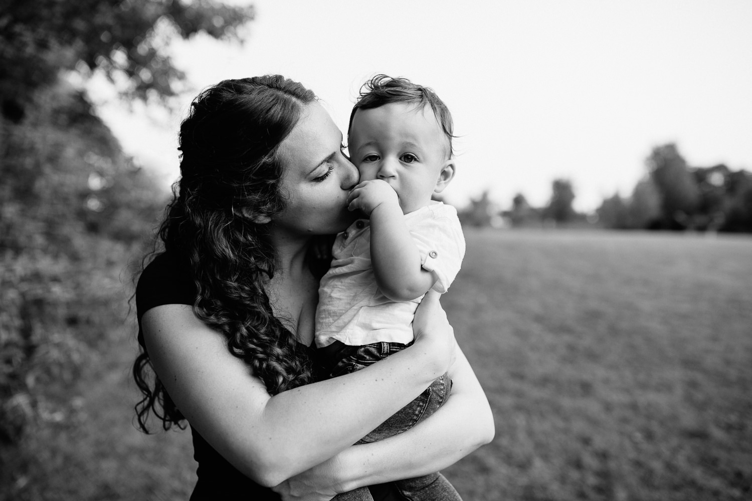 mom standing holding 9 month old baby boy with dark hair wearing white t-shirt and jeans, kissing son on cheek, boy looking at camera - GTA Lifestyle Photography
