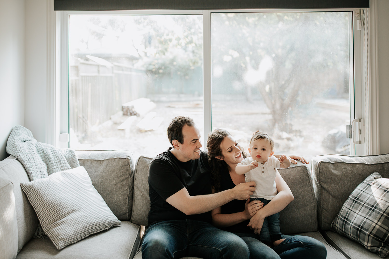 family of 3 sitting on couch, 9 month old baby boy with dark hair standing on mom's lap looking at camera, parents smiling at son - Stouffville Golden Hour Photography