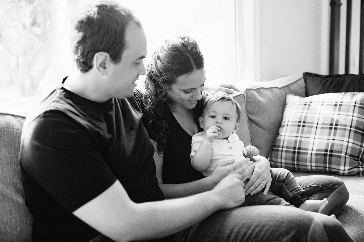 family of 3 sitting on couch, 9 month old baby boy with dark hair sitting on mom's lap looking at camera, parents smiling at son - Markham Golden Hour Photography