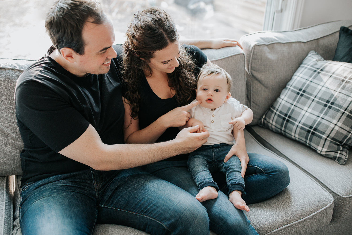 family of 3 sitting on couch, 9 month old baby boy with dark hair sitting on mom's lap looking at camera, parents smiling at son - Newmarket Golden Hour Photography