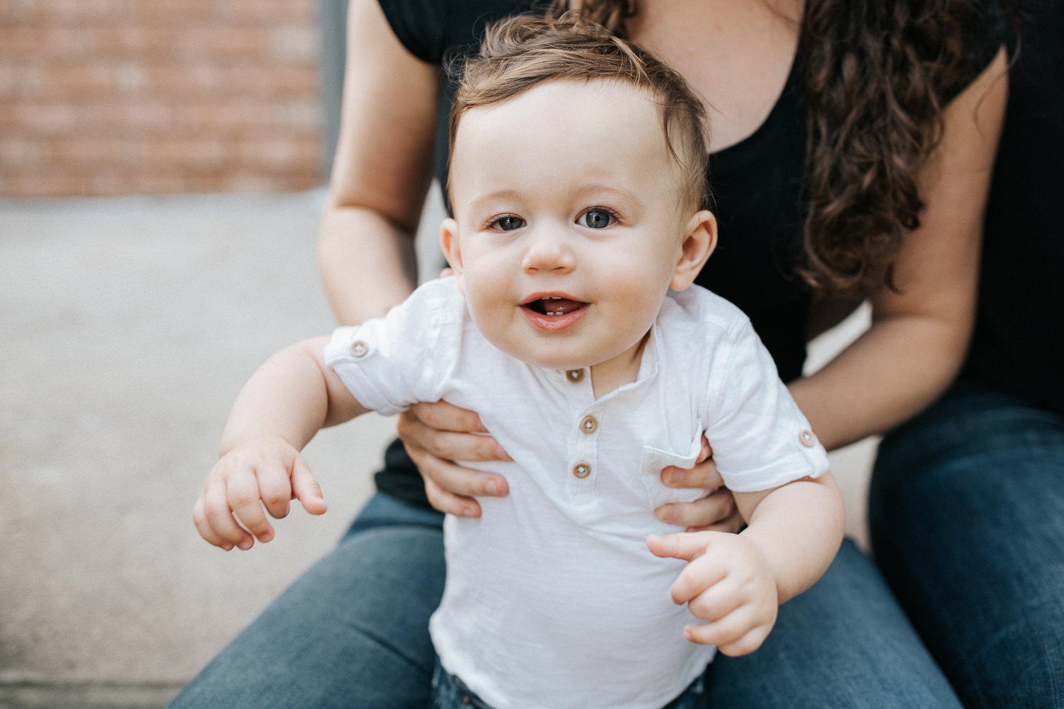 9 month old baby boy with dark hair in white t-shirt standing in mom's lap, smiling at camera - Barrie Golden Hour Photography