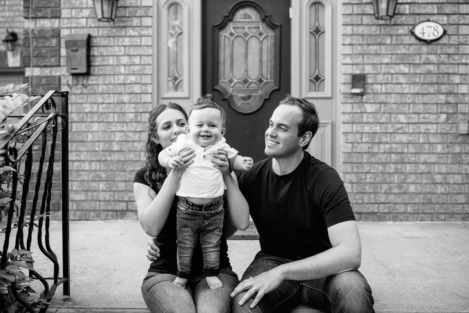 family of 3 sitting on front steps of home, mom holding 9 month old baby boy with dark hair who is standing in her lap, dad sitting next to wife and son smiling - GTA Lifestyle Photos