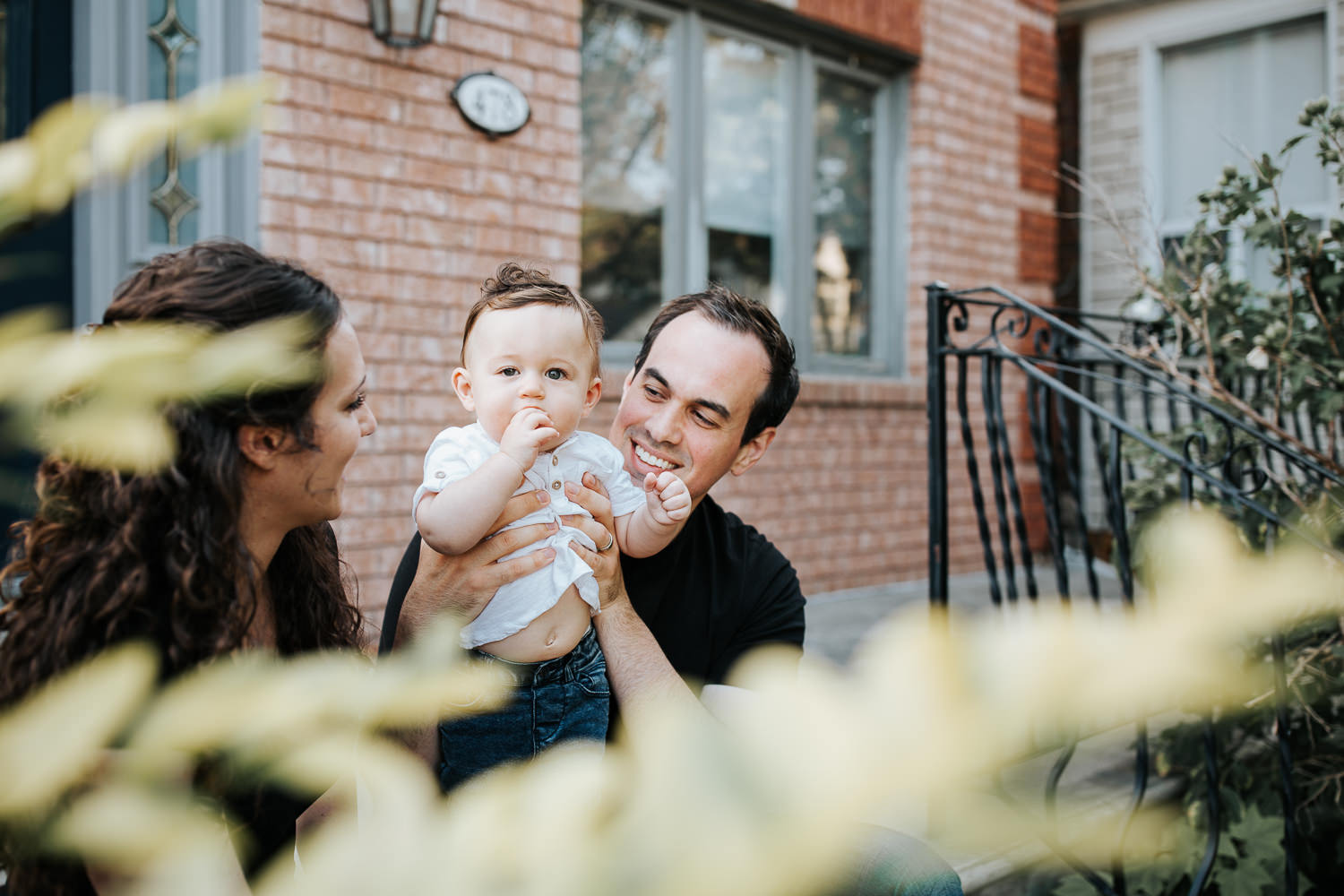 family of 3 sitting on front porch, 9 month old baby boy with dark hair in white t-shirt and jeans standing in dad's lap, mom smiling at son and husband - York Region Golden Hour Photography