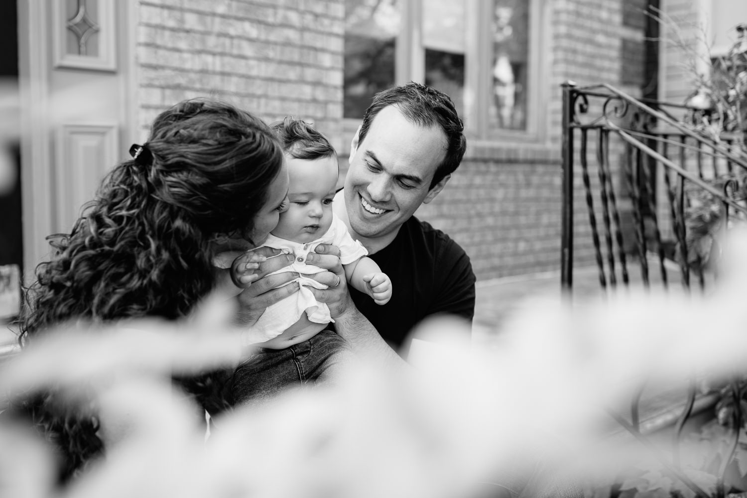 family of 3 sitting on front porch, 9 month old baby boy with dark hair in white t-shirt and jeans standing in dad's lap, mom kissing son on cheek - York Newmarket Golden Hour Photos