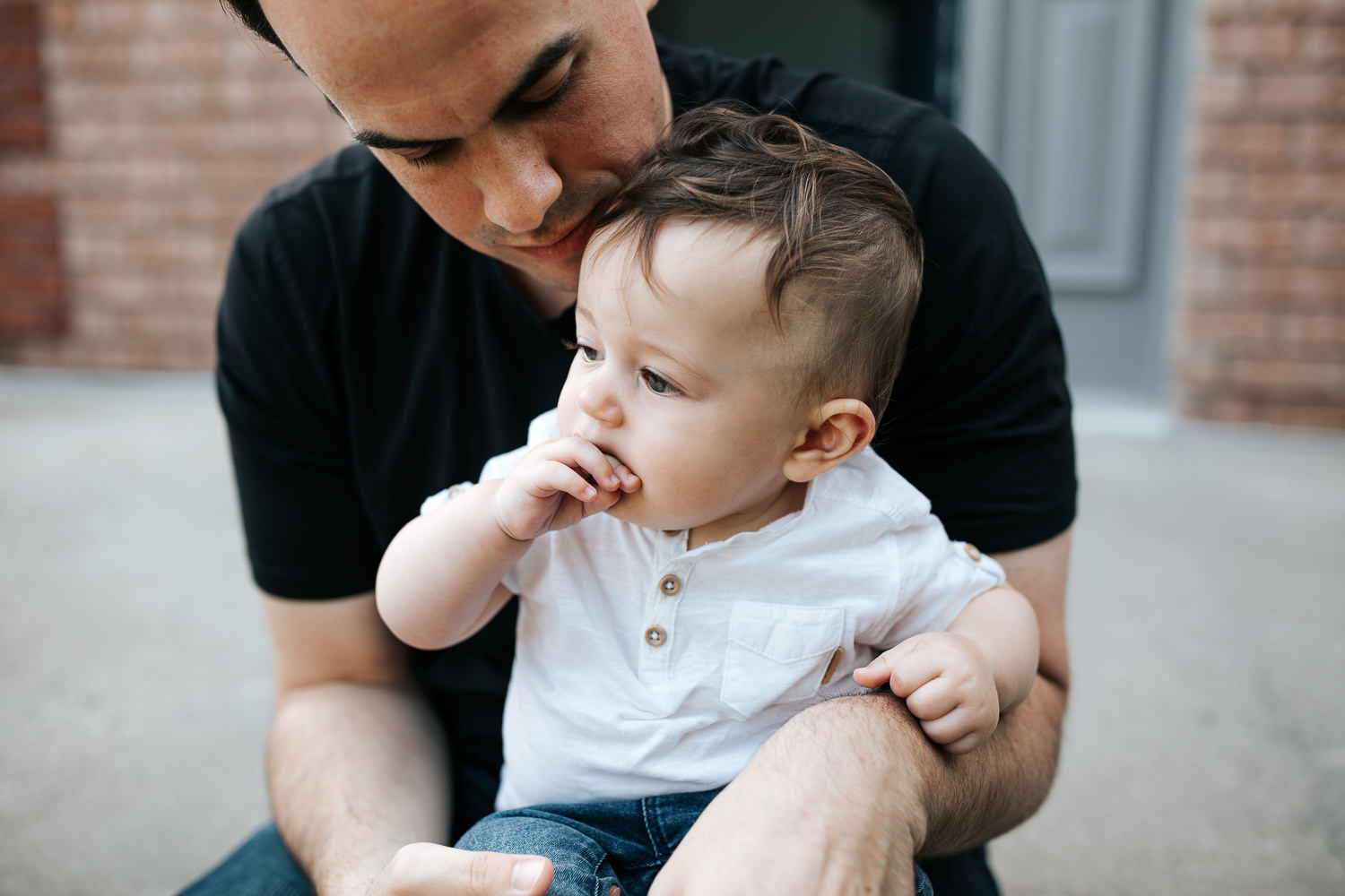 9 month old baby boy with dark hair in white t-shirt and jeans sitting in dad's lap chewing on hand - Stouffville Golden Hour Photos