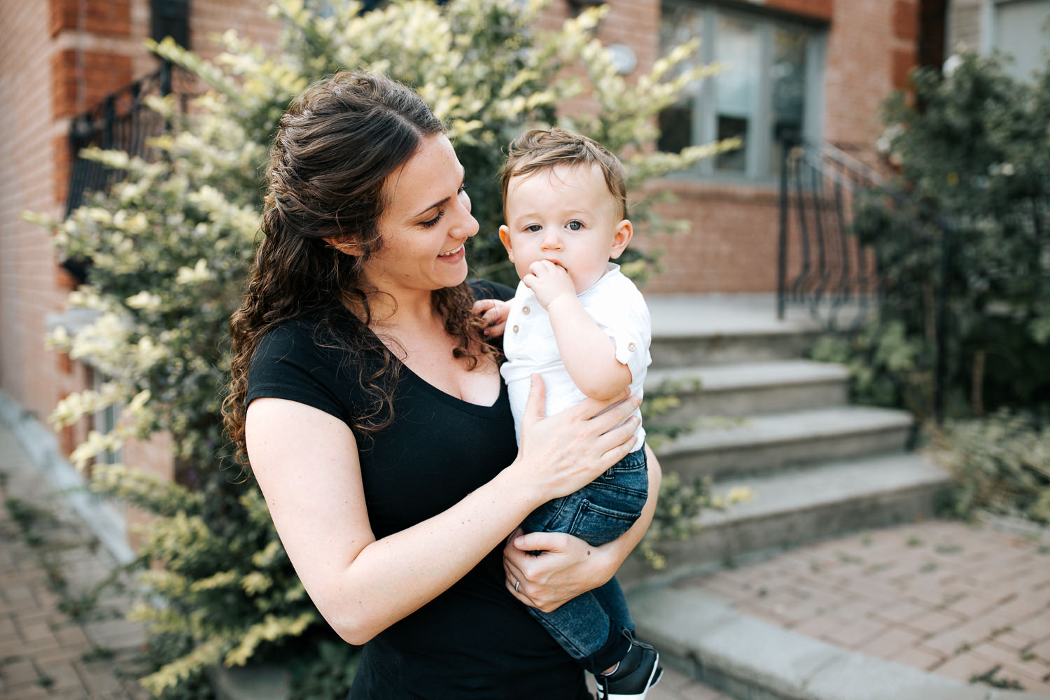 mom standing holding 9 month old baby boy with dark hair wearing white t-shirt and jeans, smiling at son, boy looking at camera - Markham Golden Hour Photos