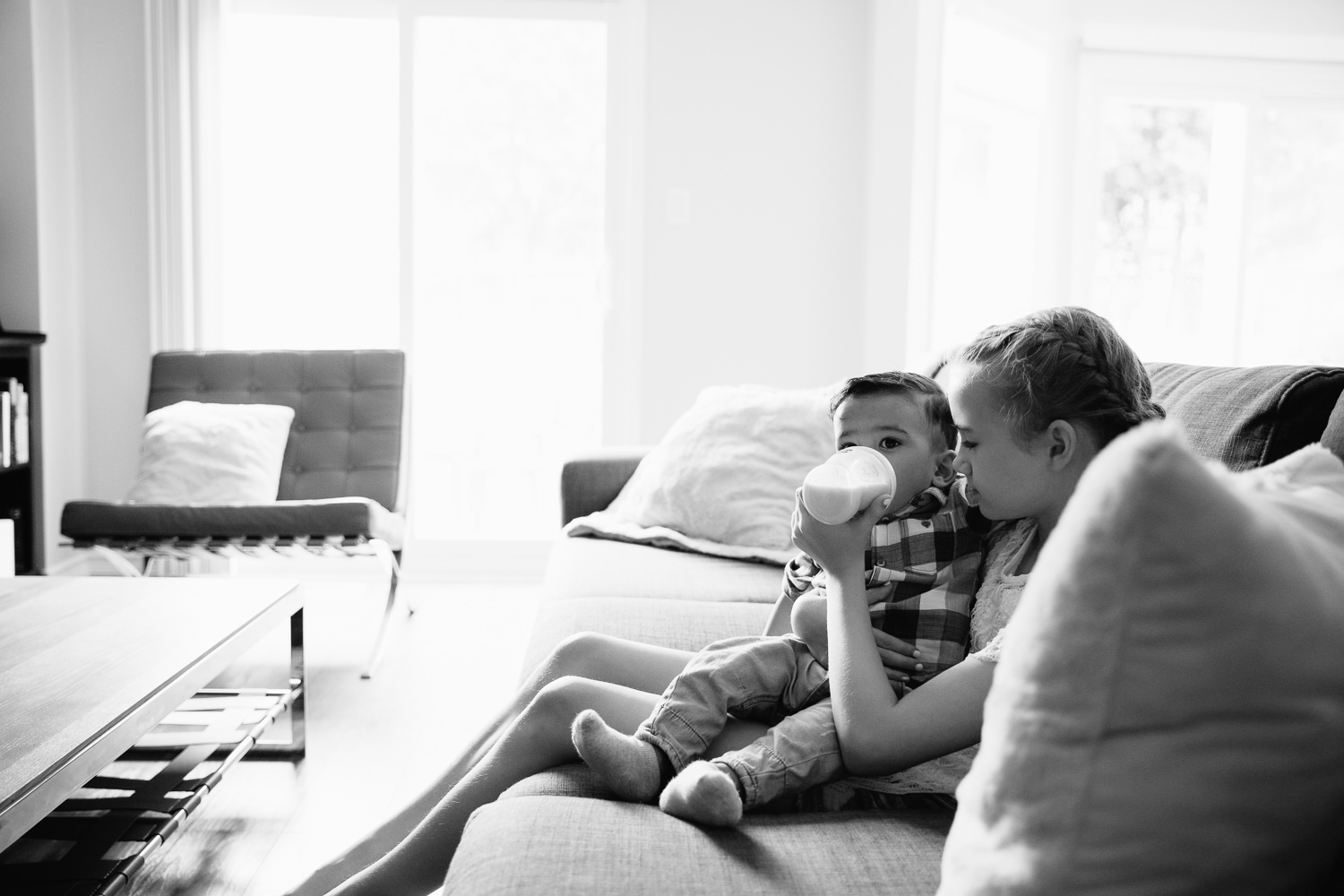 11 year old girl with blonde hair sitting on couch feeding 1 year old dark haired boy in shirt and jeans a bottle - Stouffville Lifestyle Photography