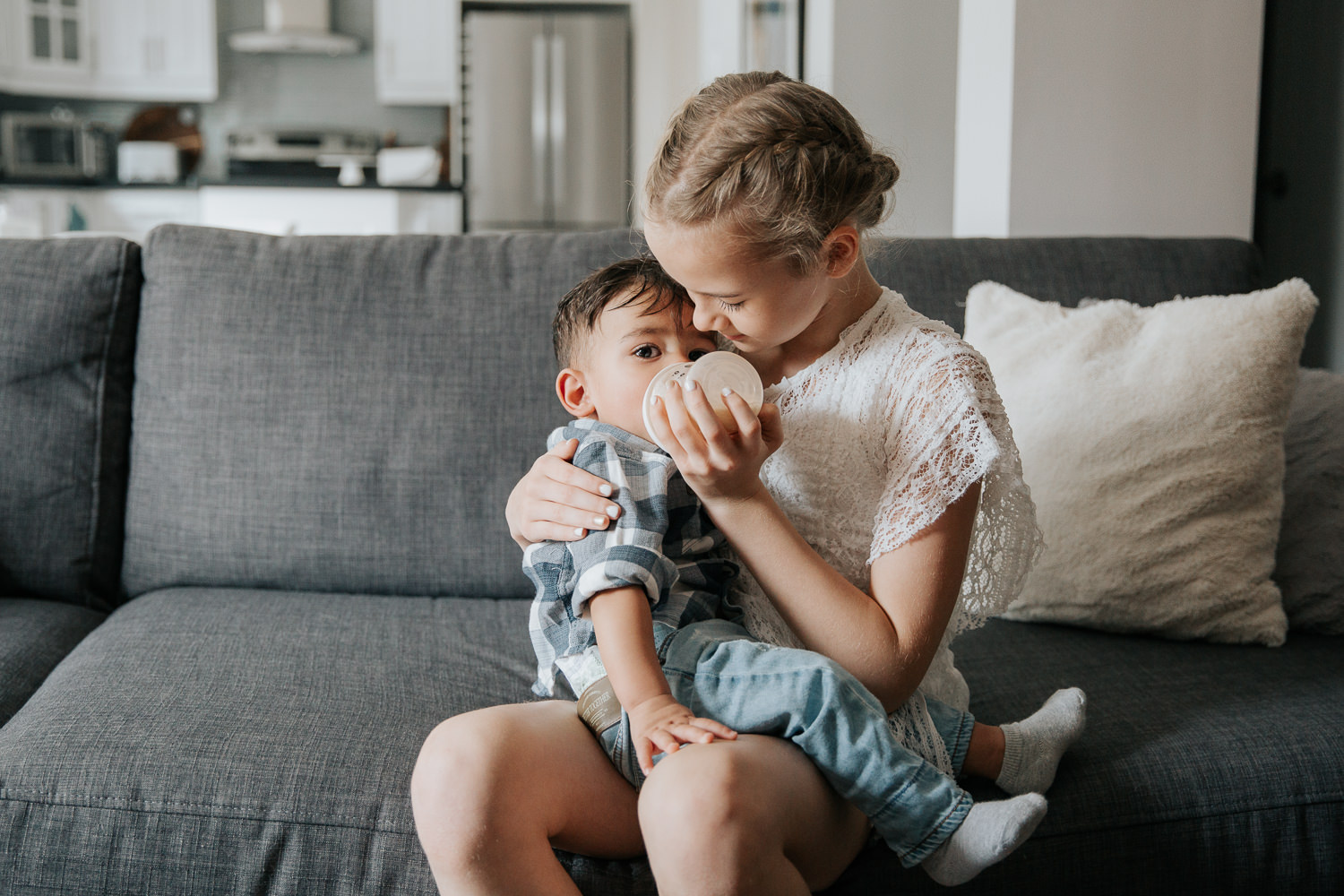 11 year old girl with blonde hair sitting on couch feeding 1 year old dark haired boy in shirt and jeans a bottle - Newmarket Lifestyle Photography