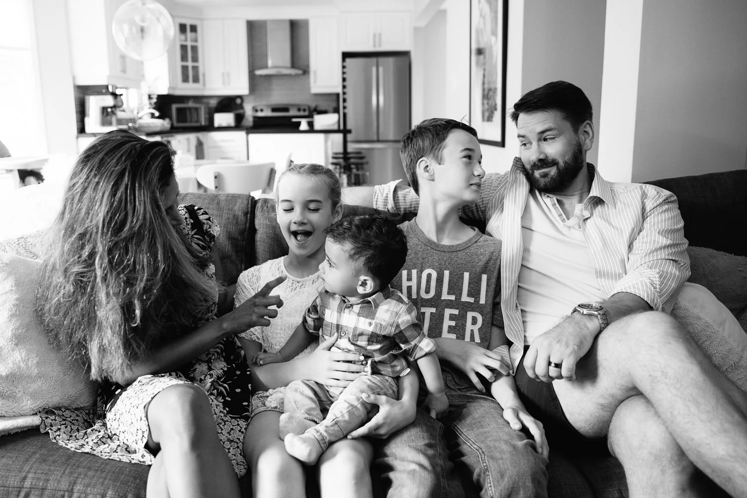 family of 5 sitting on grey couch smiling at one another, 1 year old baby boy sitting in older sister's lap, next to brother, parents on each side - Barrie Lifestyle Photography
