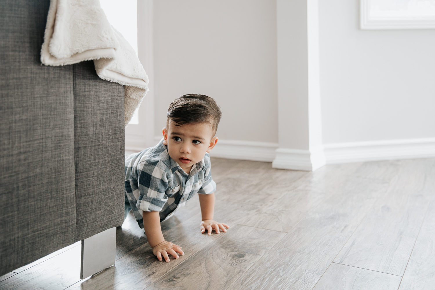 1 year old baby boy with dark hair and eyes in plaid blue and white button down shirt crawling on hardwood floor past couch - GTA Lifestyle Photography