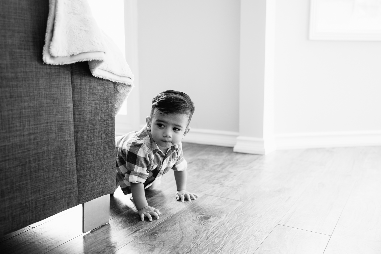 1 year old baby boy with dark hair and eyes in plaid button down shirt crawling on hardwood floor past couch - York Region Lifestyle Photography