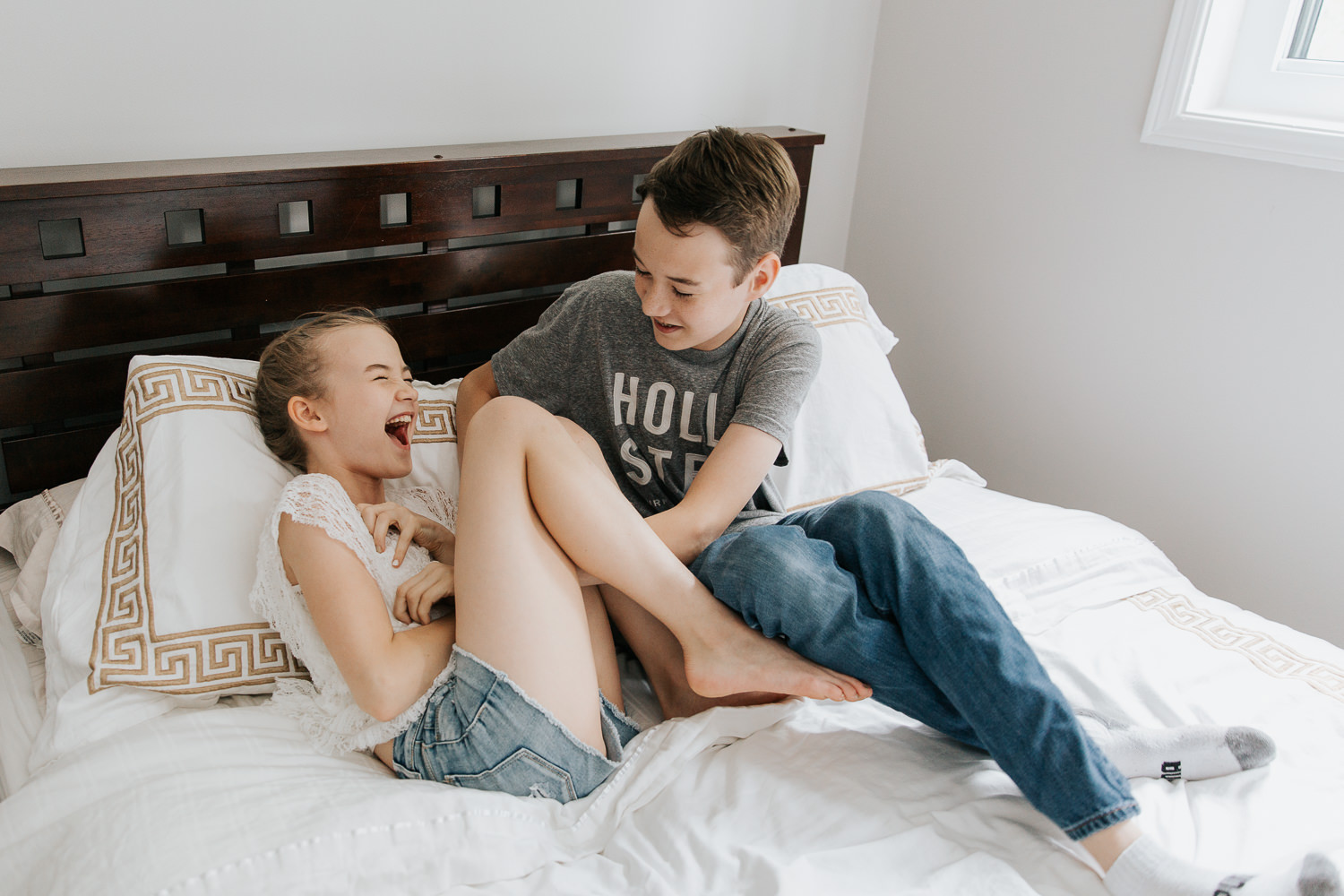 11 year old girl and 13 year old boy sitting on white bed, brother tickling sister as she laughs - Barrie In-Home Photography